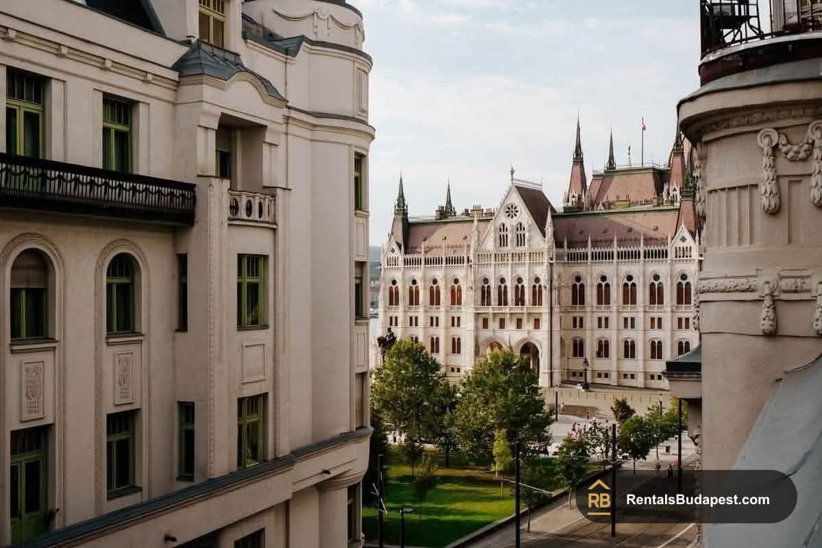 Cream neoclassical facade with green-framed windows and a balcony, overlooking the Gothic Parliament building beyond trees, Akadémia utca