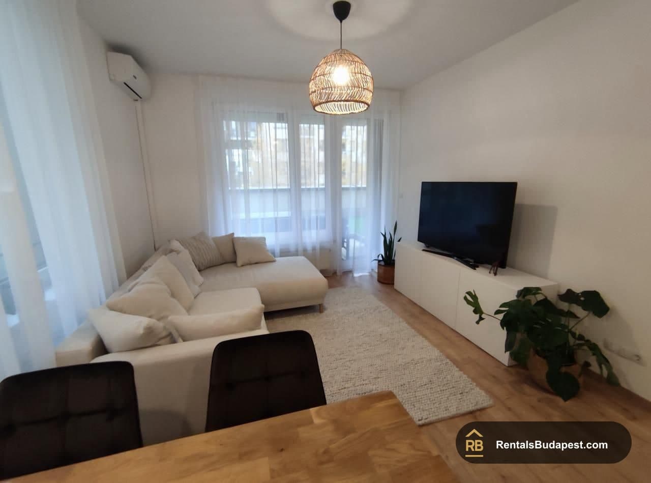 Bright living room with cream sectional sofa, wicker pendant lamp, sheer curtains, wall-mounted TV and wooden floor, Corvin sétány